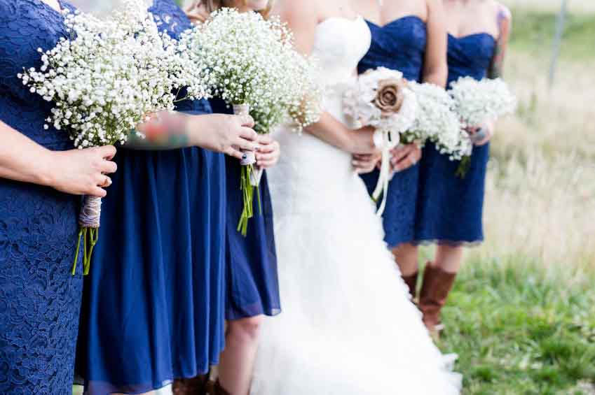 Decorative beautiful fresh wedding bouquets of colorful rose flowers pink violet lilac purple white orange and yellow in hands of bride and bridesmaid in blue dresses, horizontal picutre