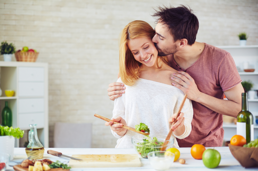 Young man kissing his wife while she mixing up salad ingredients
