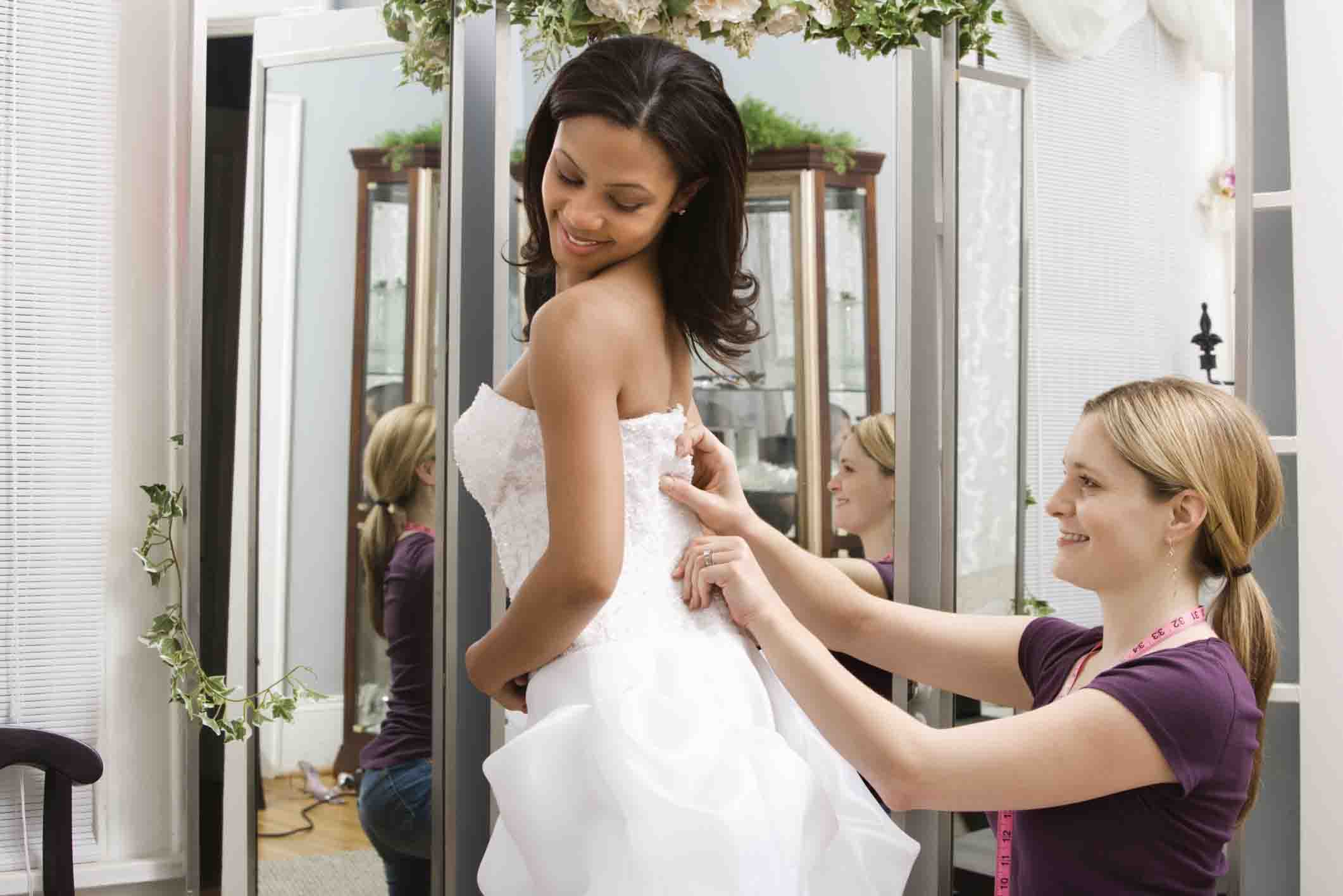 Caucasian seamstress helping African-American bride in bridal shop.