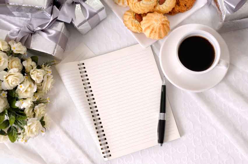 Folded notebook and pen laid on bridal lace with several silver wedding gifts and fabric rose bouquet, with cup of coffee and biscuits.