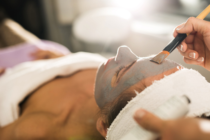 Close up of unrecognizable beautician applying clay mask on woman's face during beauty treatment.