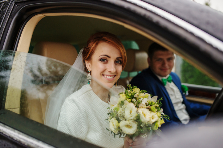 Smiled red haired bride siiting with groom at car and looking from window at rain.