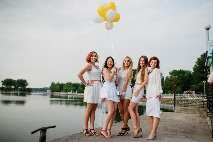 Five girls with balloons at hand weared on white dresses on hen party against pier on lake.