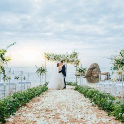 Bride and groom standing at a beachfront wedding ceremony with floral arrangements, white chairs, and the ocean in the background